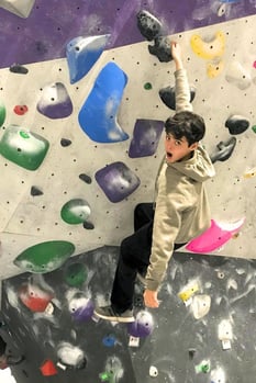A Rock Point School student hangs from a rock climbing wall, having fun and making a goofy face.