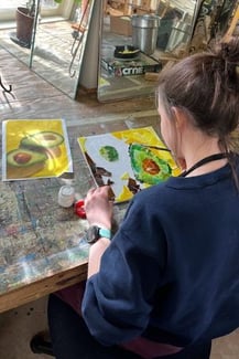 A female presenting student at Rock Point School works on an art project at school, the shot is over the students shoulder, looking down at what they're painting. the painting is of an avocado split open in half.