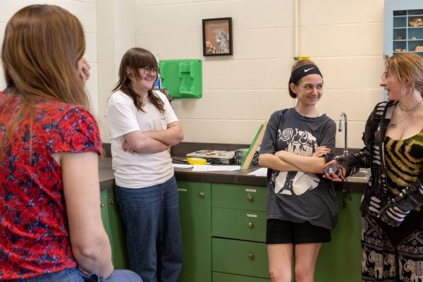 3 students stand together at the counter of the science classroom having a conversation with the teacher who is sitting with their back towards the camera. 