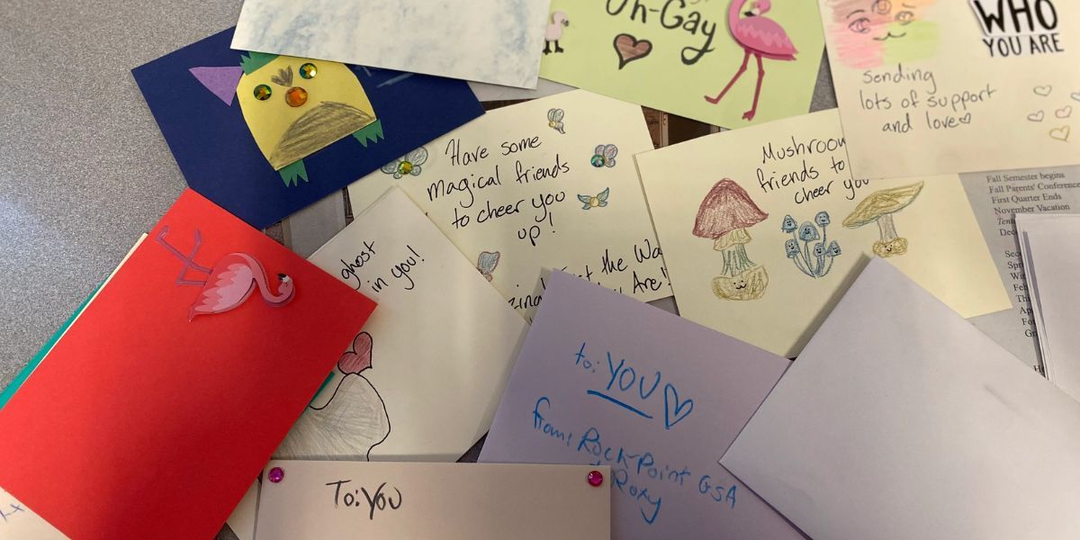 A pile of hand written and hand drawn cards are cluttered all over a table, letters and art overlapping each other. Letters are colorful, fun, and have supportive messaging to be yourself and love yourself.