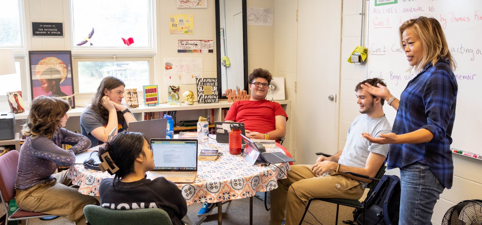 A group of students sit at a table and listen to the teacher standing by them as the teacher enthusiastically talks. 
